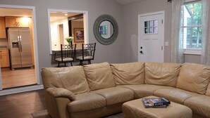 A tan leather sectional sofa, a dining table with chairs, a kitchen area, and a door with a window view. - The Foxboro Game Day House — 7 Min to Gillette (Foxborough)