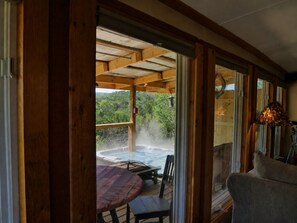 A wooden deck with a hot tub, a table, and chairs, overlooking a lush green landscape.