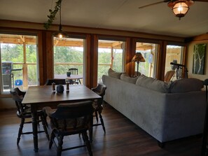 A wooden dining table with chairs, a sofa, and a ceiling fan in a room with large windows and wooden beams.
