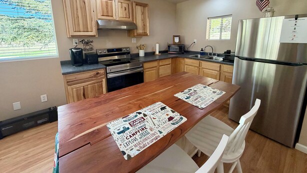 A modern kitchen with wooden cabinets, stainless steel appliances, a wooden dining table with placemats, and a view of the outdoors. - The Lookout at Silver Springs! Mountain air and Trout Pond Fun-sleeps up to 5 (Cloudcroft)