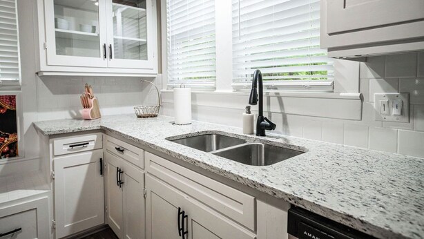 Una cocina con armarios blancos, encimera de granito, pileta doble y ventana con persianas.