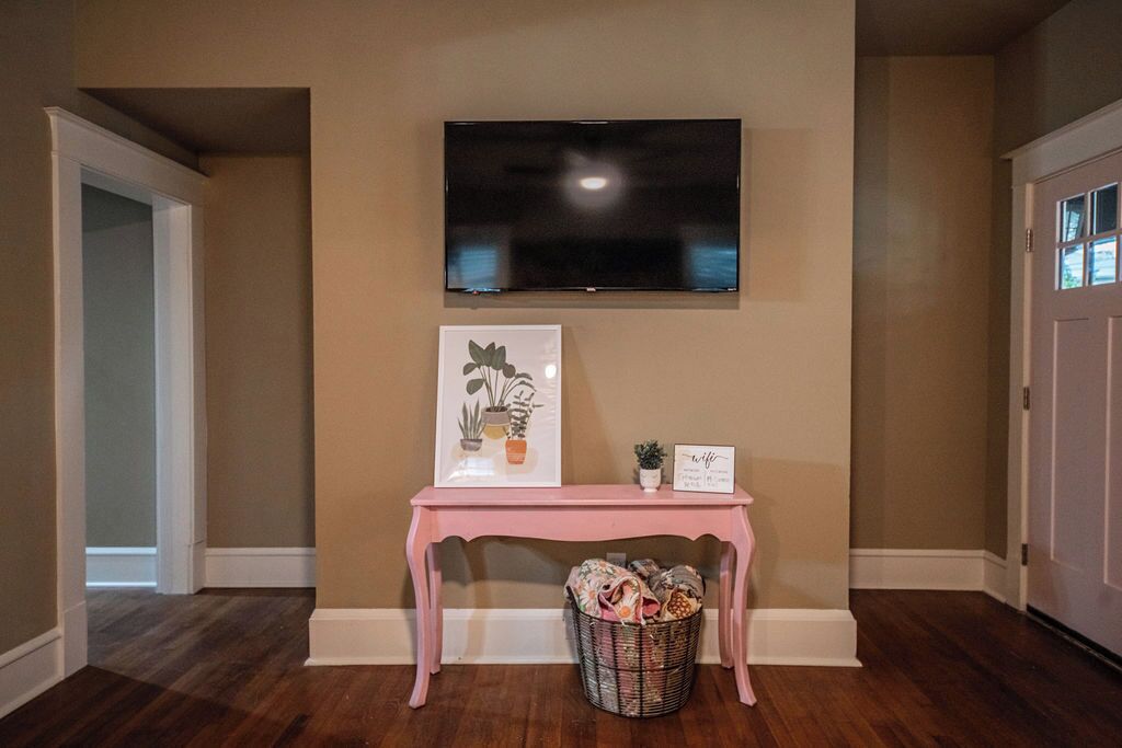 Une pièce avec un téléviseur fixé au mur, une console rose sur laquelle est posé un panier, et un tableau représentant des plantes.