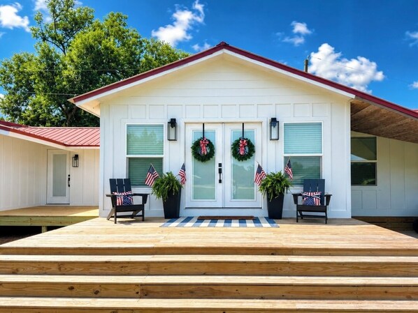 A white house with a red roof, two front doors, and a porch with two chairs and American flags.