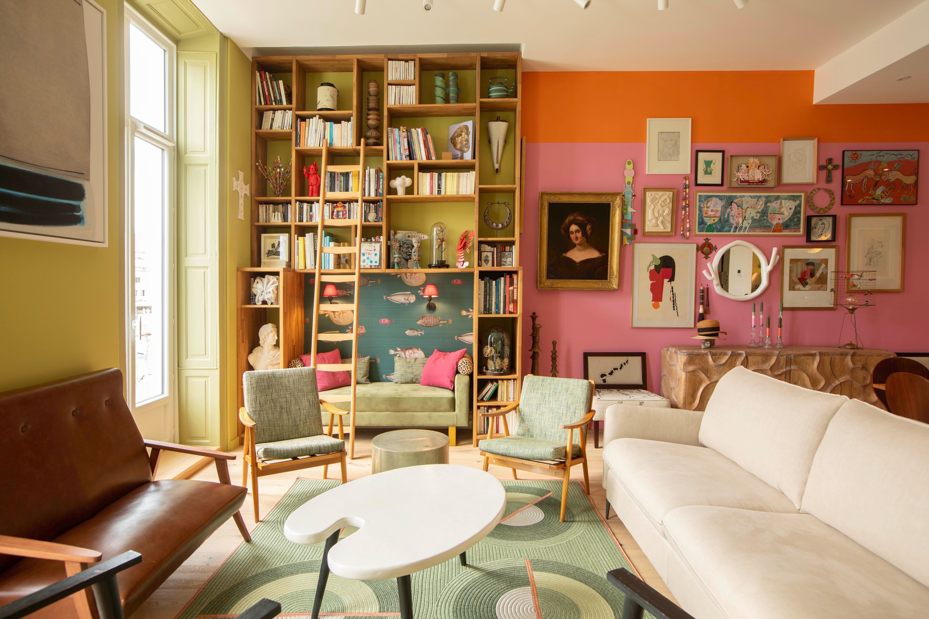 Un salon chaleureux avec un canapé vert, deux fauteuils, une table basse blanche et une bibliothèque remplie de livres et d’objets décoratifs.