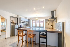 A modern kitchen with a central island, two wooden bar stools, a built-in oven, and a window with heart-shaped decorations. - Le Col de Feu - Joli duplex pour 8 avec jardin (Lullin)