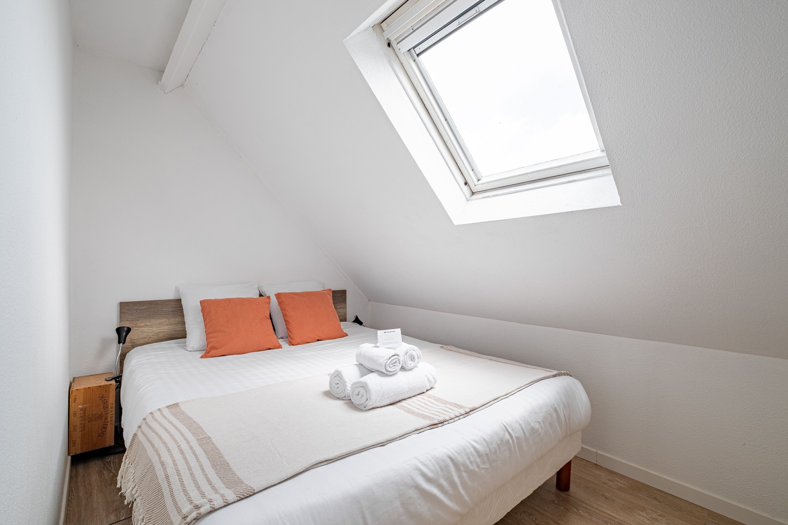 A bedroom with a sloped ceiling, a bed with white bedding and orange pillows, a wooden nightstand, and a window.