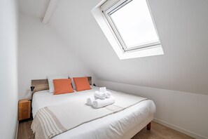 A bedroom with a sloped ceiling, a bed with white bedding and orange pillows, a wooden nightstand, and a window. - Les 4 Voiles - À 280 m de la plage (Damgan)