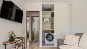 Een ruimtelijk ingerichte leefruimte met een flatscreen-tv, een wasmachine, een bank en een console tafel met boeken en een vaas.