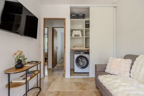 A compact living space with a flat-screen TV, a washing machine, a sofa, and a console table with books and a vase. - Les Oliviers du Papet - Le Nid (Châteaurenard)
