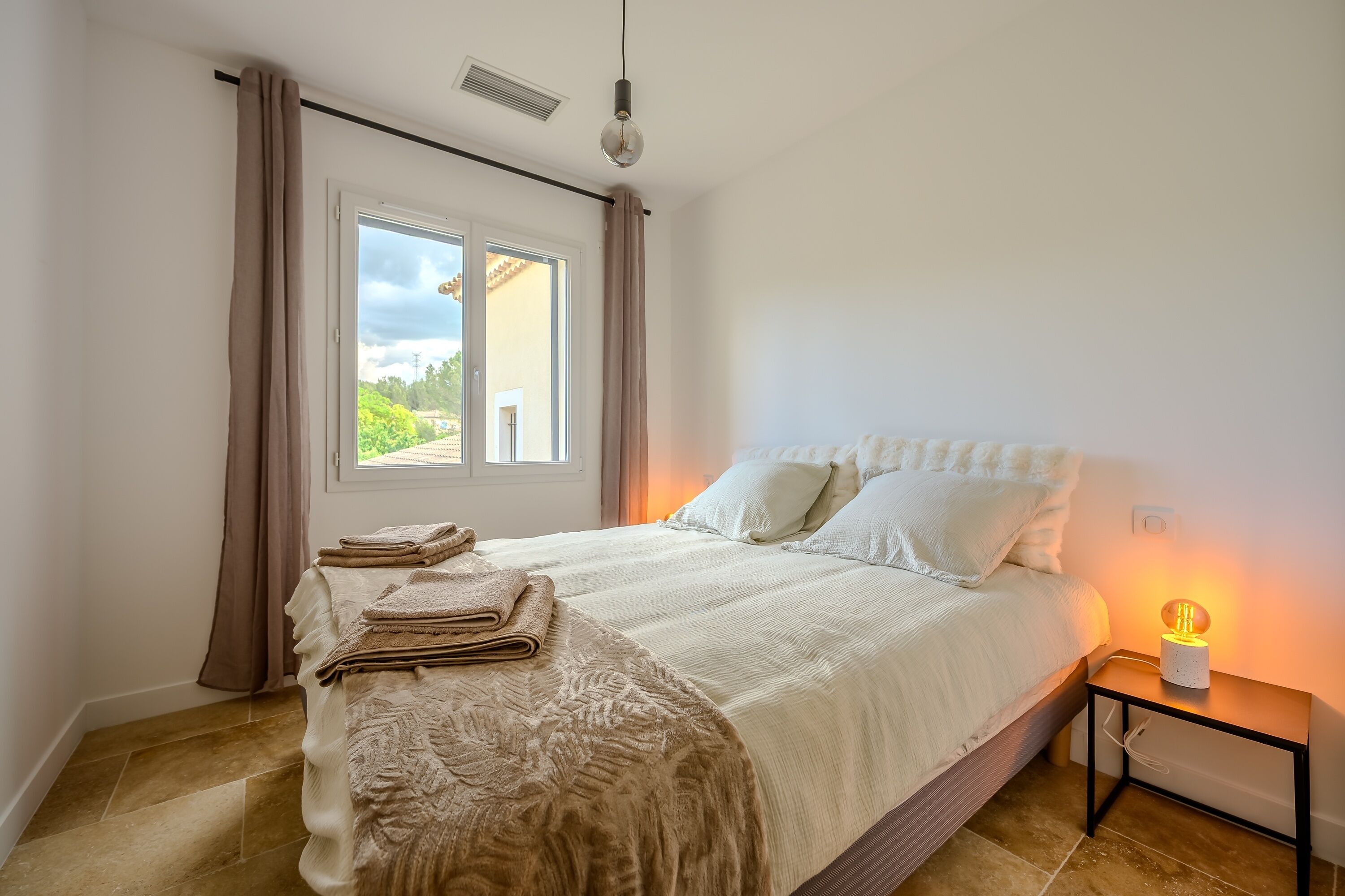Un dormitorio con una cama, una mesita de noche con lámpara, una ventana con cortinas y vistas al exterior.