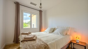 A bedroom with a bed, a nightstand with a lamp, a window with curtains, and a view of greenery outside. - Les Oliviers du Papet - Le Nid (Châteaurenard)