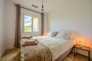 A bedroom with a bed, a nightstand with a lamp, a window with curtains, and a view of greenery outside. - Les Oliviers du Papet - Le Nid (Châteaurenard)