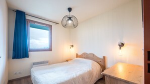 A bedroom with a bed, a window with curtains, a wooden headboard, a bedside table, and a wall-mounted light fixture. - Les Alpages du Corbier B108 - Télésièges à 250 m (Villarembert)