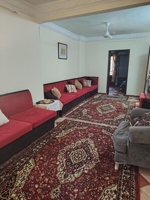 A living room with a red sofa, a grey armchair, a patterned carpet, and a ceiling fan. - Apartment in very quiet area 5 mn from piramides, (Guiza)
