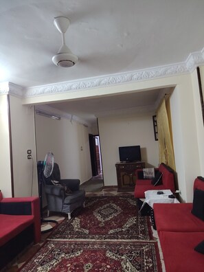 A living room with a ceiling fan, a red sofa, a black armchair, a television, and a patterned rug. - Apartment in very quiet area 5 mn from piramides, (Guiza)