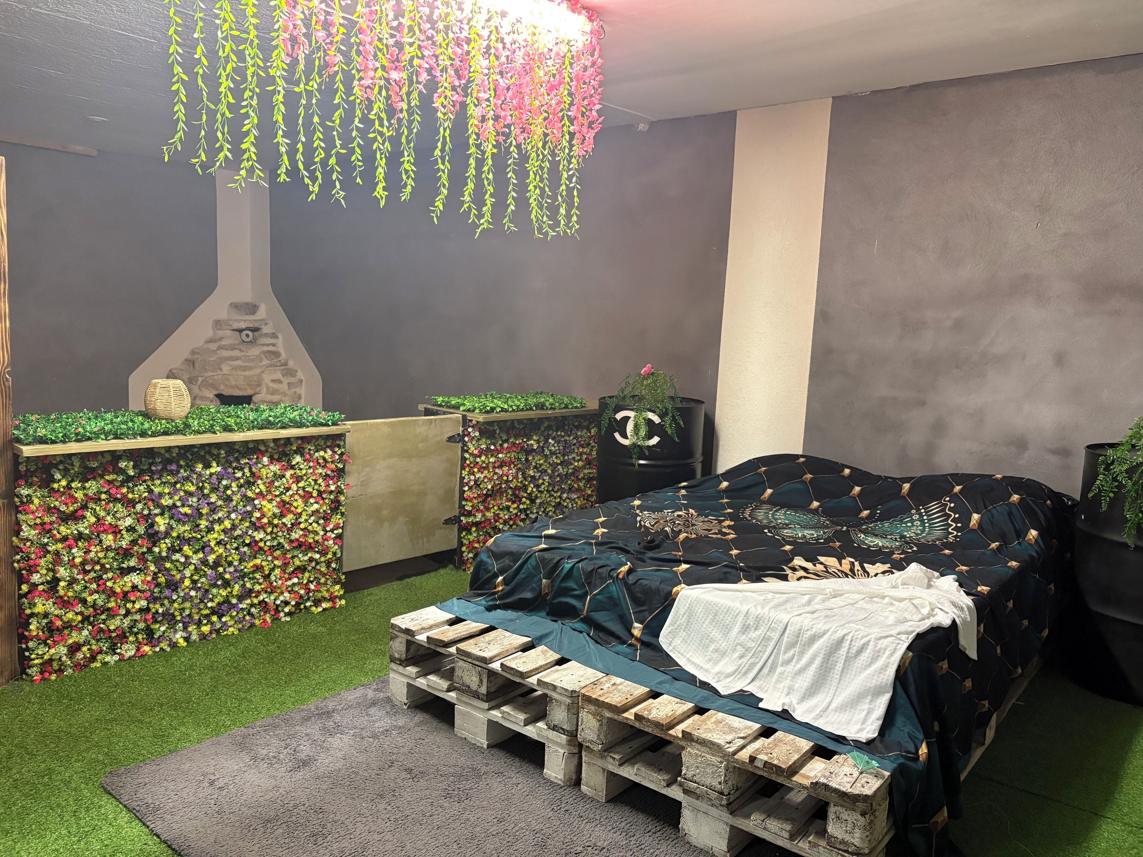A bedroom with a bed, a wooden pallet bench, a stone fireplace, and a wall of artificial flowers.
