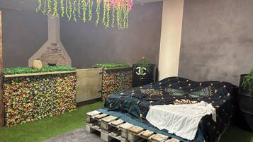 A bedroom with a bed, a wooden pallet bench, a stone fireplace, and a wall of artificial flowers.