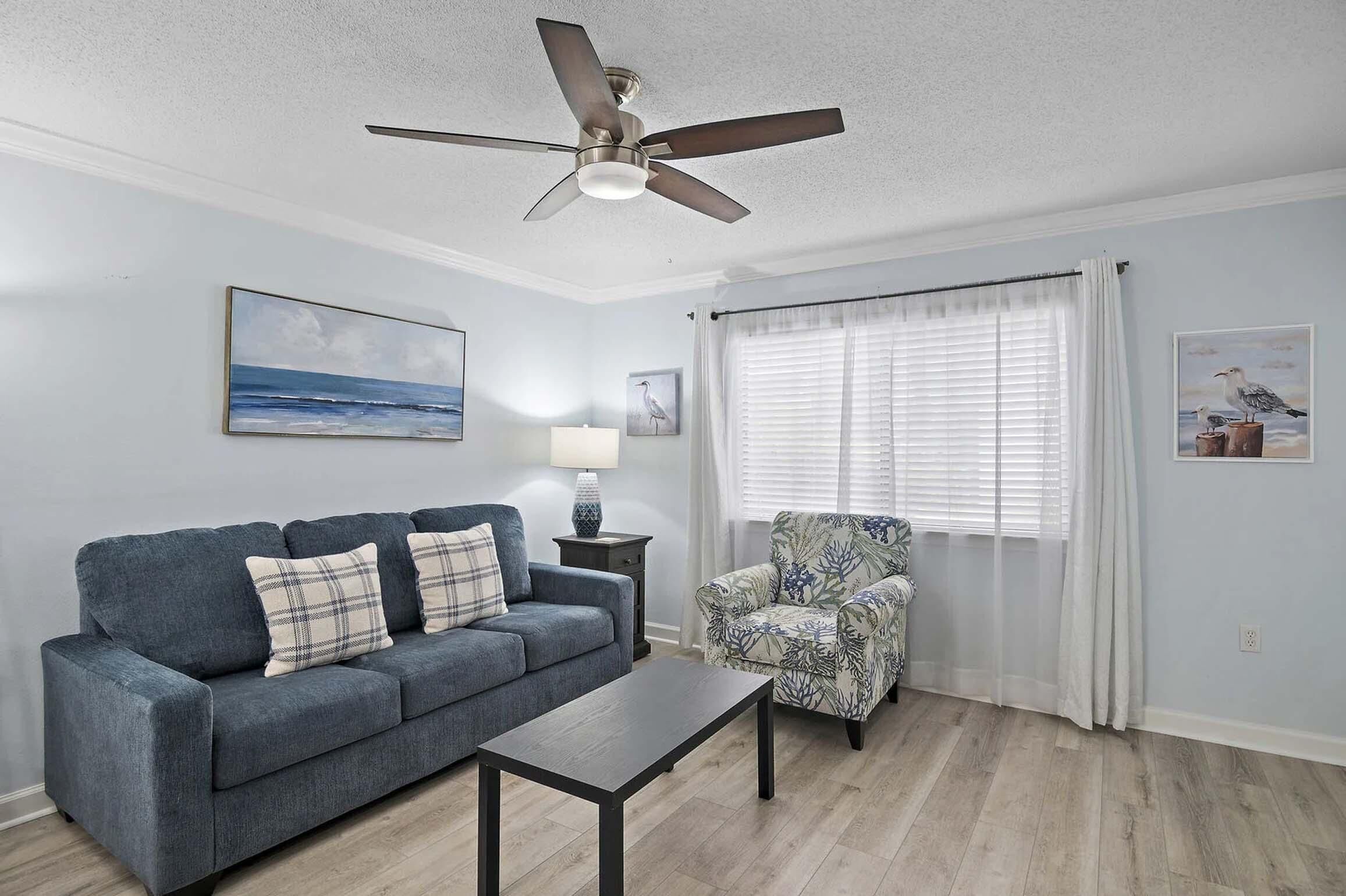 A living room with a sofa, armchair, coffee table, ceiling fan, and artwork on the walls.