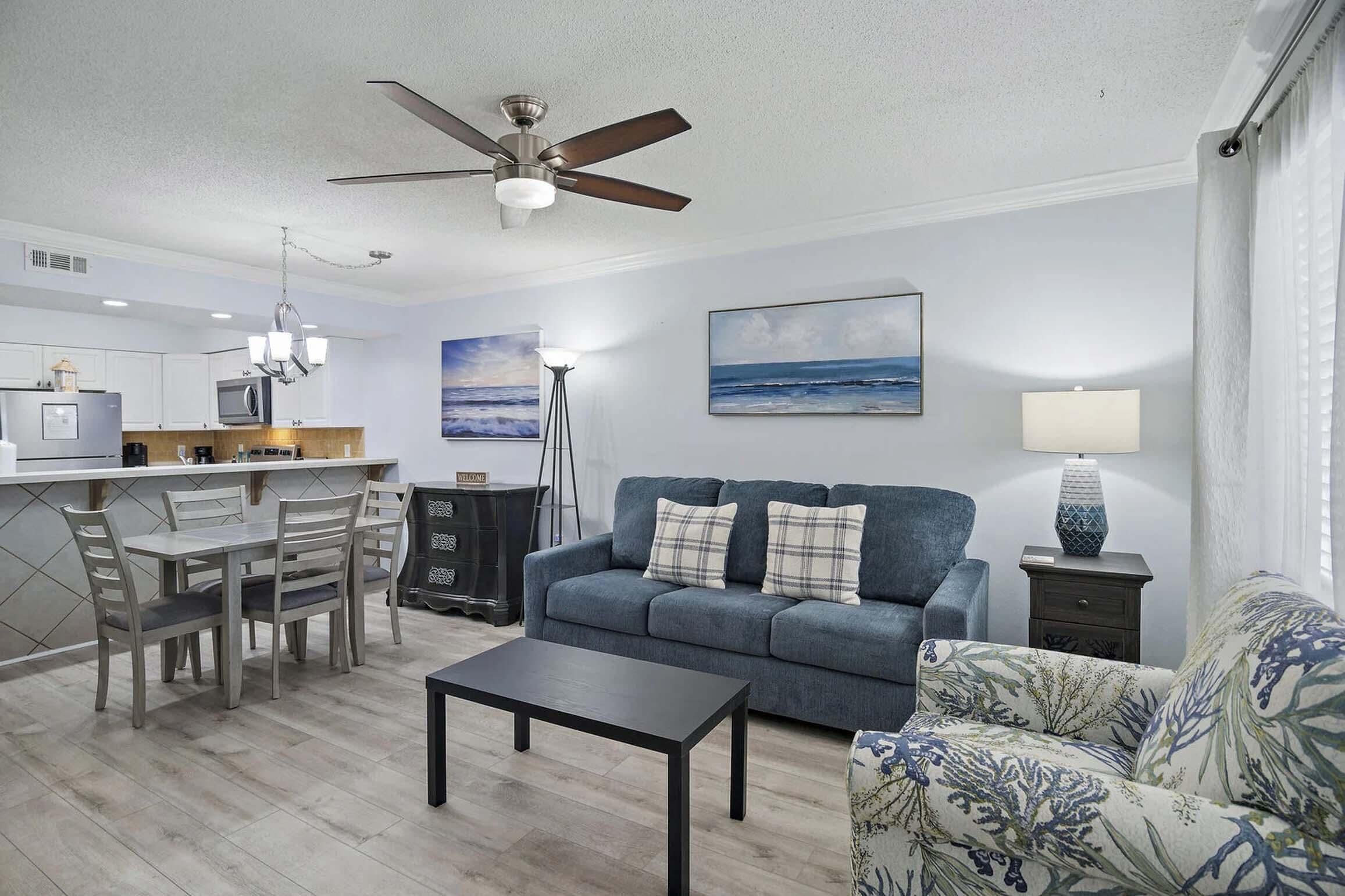 A modern living room with a sofa, a coffee table, a dining area with chairs, a kitchen with a fridge, and a ceiling fan.