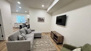 Un salon moderne avec un canapé d’angle gris, un fauteuil vert, un tapis à motifs, un meuble TV en bois et, au fond, un coin repas.