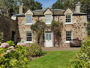 A stone building with a central door, two windows on each side, and a small garden area with flowers and a bench.