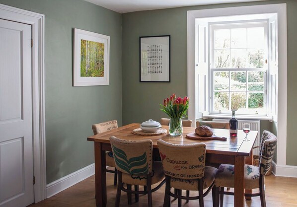 A dining area with a wooden table, chairs, a vase with flowers, and a window with a view of greenery.