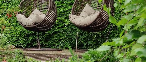 Two wicker hanging chairs with cushions against a wall of dense green foliage.