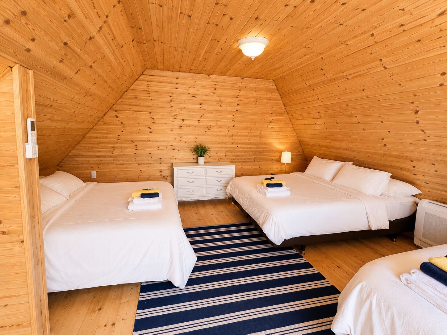 Un cuarto acogedor y triangular, con paredes de madera y techo inclinado. Hay dos camas con sábanas blancas, un pequeño tocador blanco y una lámpara sobre él.