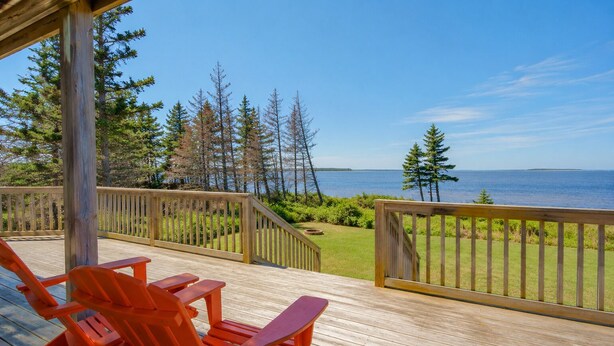 Eine Holzterrasse mit orangefarbenen Stühlen, Blick aufs Meer und eine Rasenfläche mit Bäumen.