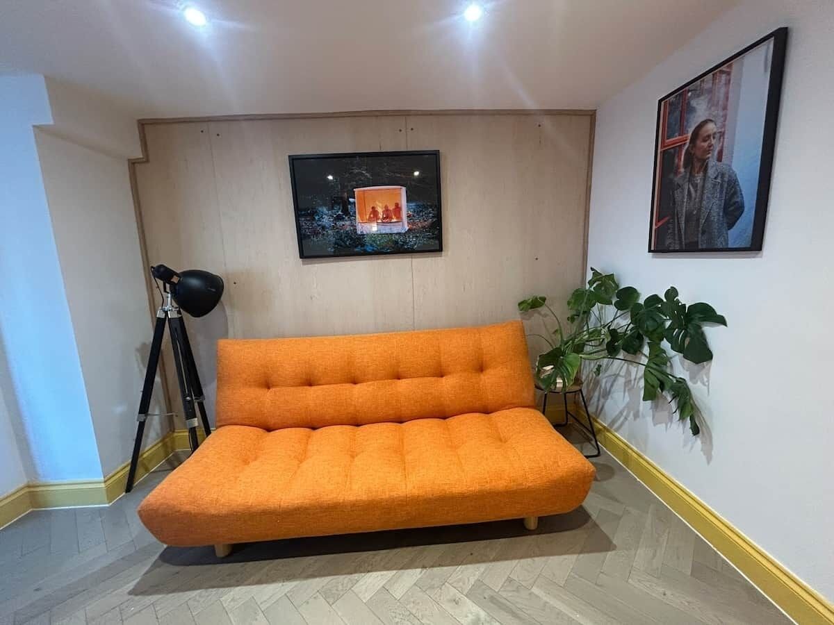 Una habitación con un sofá naranja grande, una lámpara con trípode, un cuadro en la pared y una planta en maceta.