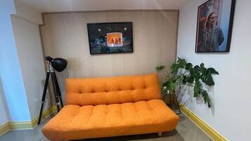 Una habitación con un sofá naranja grande, una lámpara con trípode, un cuadro en la pared y una planta en maceta.