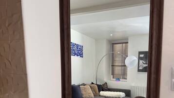 Een houten console tafel met gouden Boeddha beeldjes, een wereldbol en een spiegel die een woonkamer weerspiegelt met een grijze bank, kussens en een raam met jaloezieën.