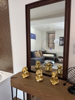 Ein Holzkonsolentisch mit goldenen Buddha-Statuen, einem Globus und einem Spiegel, der ein Wohnzimmer mit grauem Sofa, Kissen und einem Fenster mit Jalousien reflektiert.