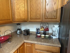 Una cocina con armarios de madera, una licuadora, un tostador, una cafetera y un estante metálico con tazas.