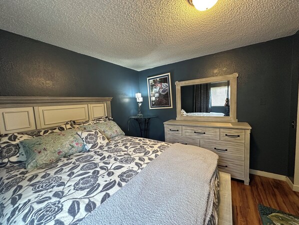 Un dormitorio con cama, cómoda, espejo, lámpara y un cuadro en la pared.