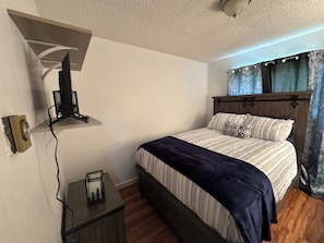 A bedroom with a bed, a TV mounted on the wall, a small table with a lamp, and a window with curtains.