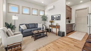 A living room with a sofa, coffee table, and a small side table with a plant. There is a kitchen area with cabinets and a refrigerator. - Henry`s Fork Suites Suite Two (Saint Anthony)