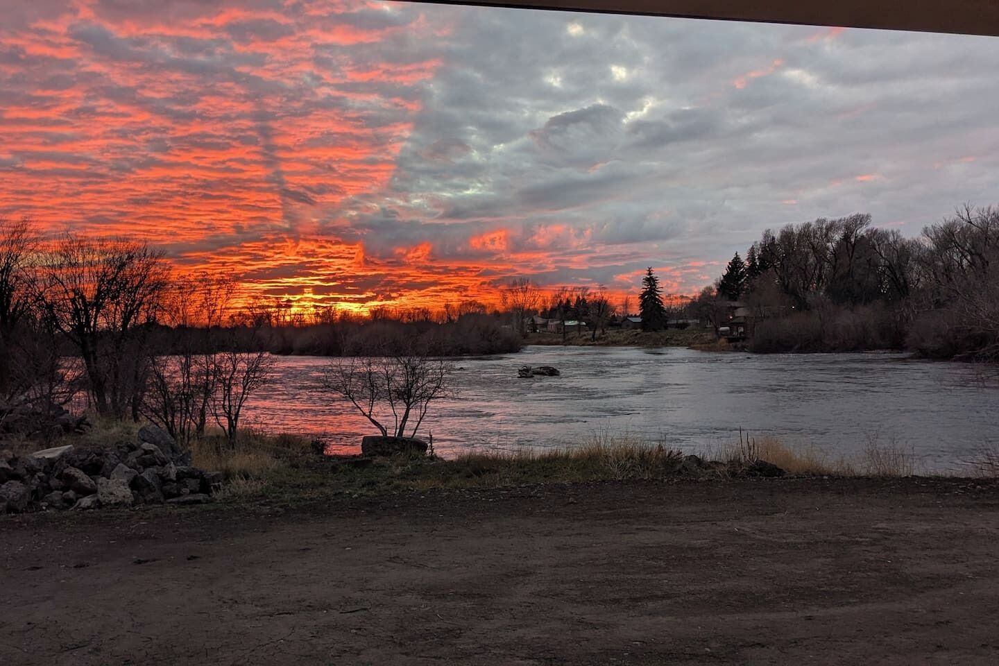 Een rivier met een ondergaande zon, omringd door bomen en enkele gebouwen.