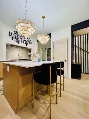 Une cuisine moderne avec un îlot central, deux chaises noires, un lustre et un escalier en arrière-plan.