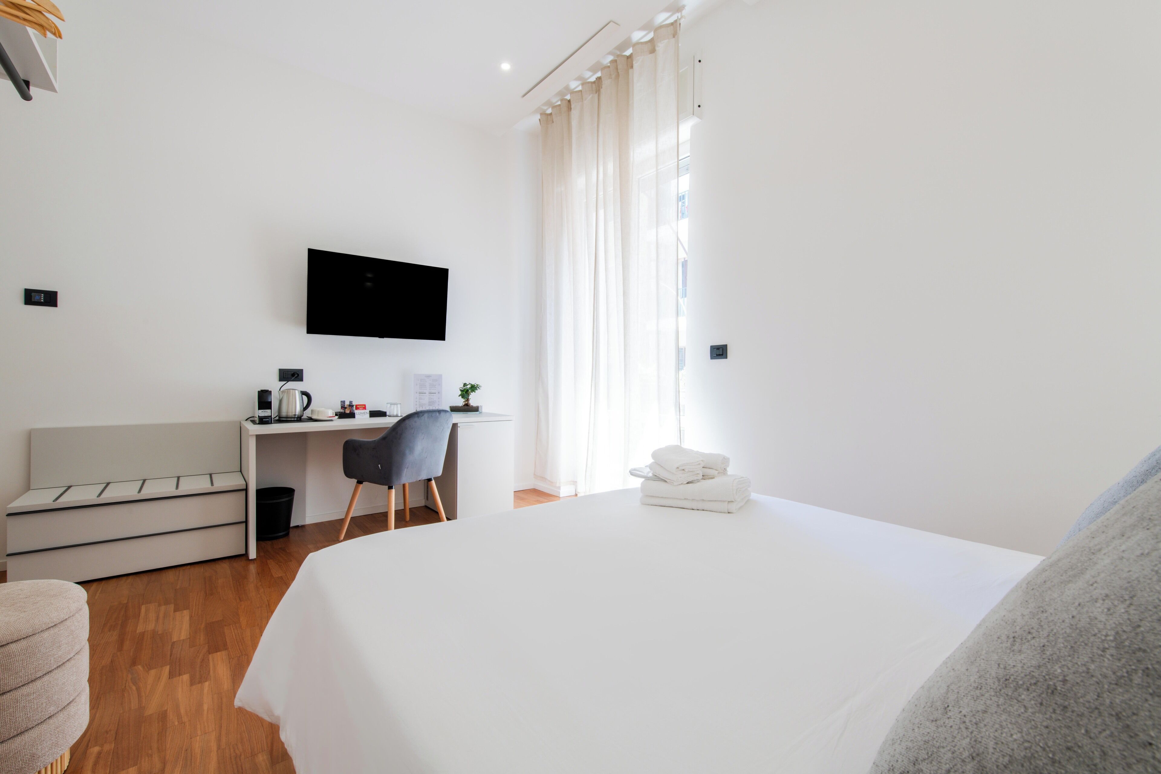 A hotel room with a bed, a desk with a chair, a television mounted on the wall, and a window with curtains.