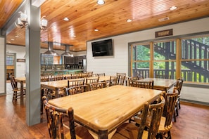 Uma sala de jantar espaçosa com mesas e cadeiras de madeira, uma cozinha ao fundo e uma grande janela com vista para árvores.