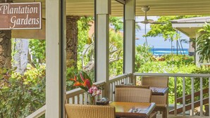 A covered outdoor seating area with wicker furniture, a sign reading 'Plantation Gardens,' and a view of the ocean. - Kiahuna 183 AC, Ground Floor Ocean View PBAC (Koloa)
