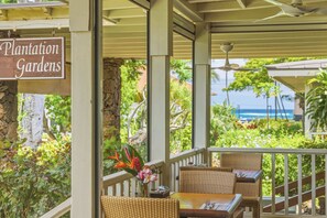 A covered outdoor seating area with wicker furniture, a sign reading 'Plantation Gardens,' and a view of the ocean. - Kiahuna 183 Ground Floor Ocean View PBAC (Koloa)