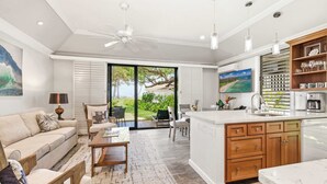 A modern kitchen with a large island, a dining area with a sofa and chairs, and a view of the outdoors through glass doors. - Kiahuna 183 AC, Ground Floor Ocean View PBAC (Koloa)