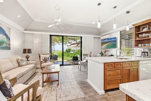 A modern kitchen with a large island, a dining area with a sofa and chairs, and a view of the outdoors through glass doors. - Kiahuna 183 Ground Floor Ocean View PBAC (Koloa)