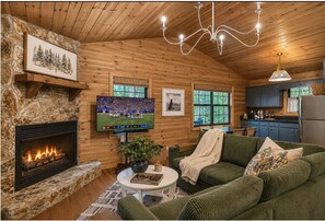 A cozy cabin interior with a stone fireplace, a television, a green sofa, and a kitchen area. - NEW Clover Ridge Cabin ~ Hot Tub + Outdoor TV +Private +Professionally Decorated (Blue Ridge)