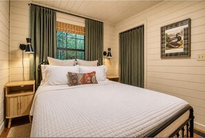 A bedroom with a bed, two bedside tables, a window with curtains, a wall-mounted lamp, and a framed picture. - NEW Clover Ridge Cabin ~ Hot Tub + Outdoor TV +Private +Professionally Decorated (Blue Ridge)