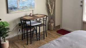 Ein Schlafzimmer mit einem Bett, einem Schreibtisch mit Stuhl, einem Bild einer Frau am Meer, einer Vase mit Trockenpflanzen und einer Tür mit einem 'welcome'-Schild.