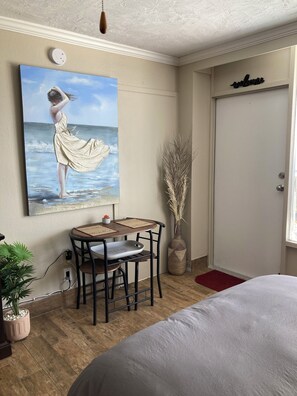 A bedroom with a bed, a desk with a chair, a painting of a woman by the sea, a vase with dried plants, and a door with a 'welcome' sign. - Cozy Quiet Beachfront Studio with Awesome Ocean View (Daytona Beach)
