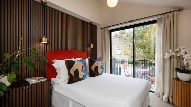 Ein Schlafzimmer mit einem großen Bett, einer Wand aus Holzpaneelen, einem roten Kopfteil, zwei Nachttischlampen, einer Topfpflanze und Blick auf einen Balkon mit Gartenmöbeln.